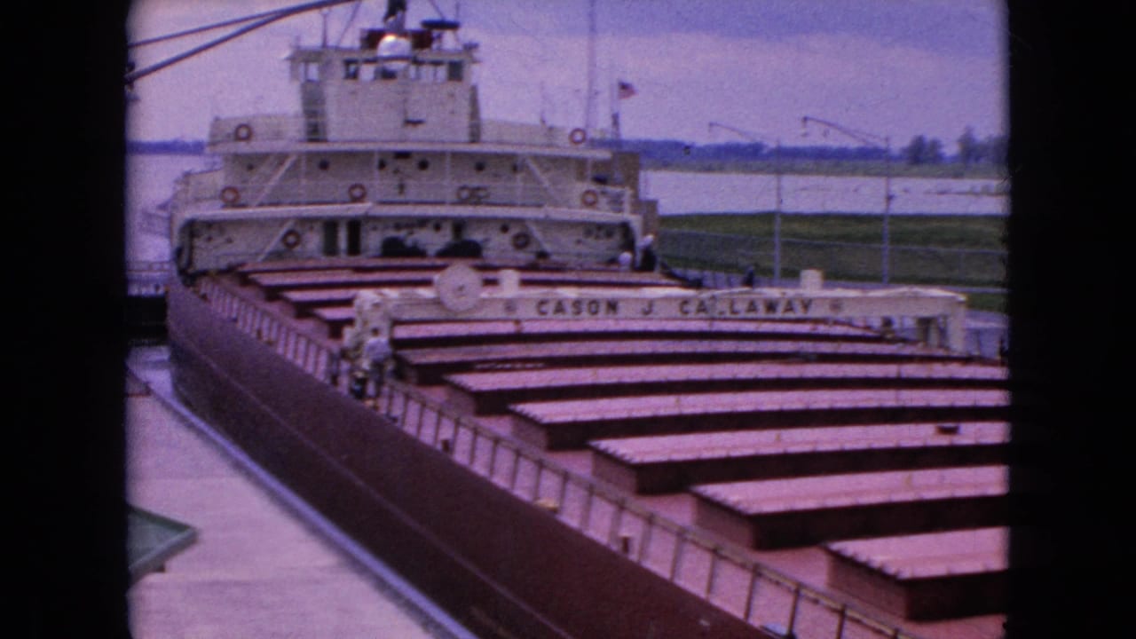 1964 8mm frame showing the docked Cason J. Callaway freighter near Niagara Falls, New York, with its white superstructure and long rows of cargo hatch covers seen through dark framing.