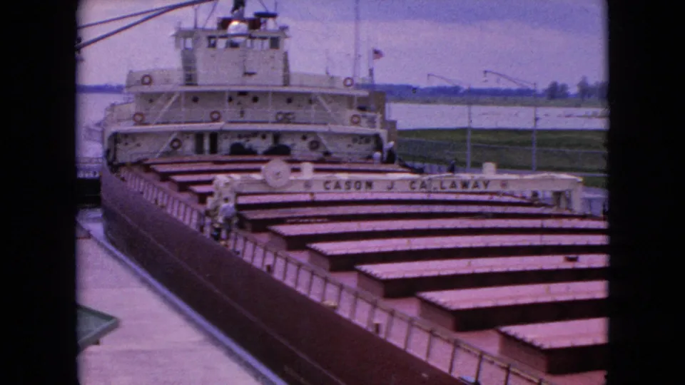1964 8mm frame showing the docked Cason J. Callaway freighter near Niagara Falls, New York, with its white superstructure and long rows of cargo hatch covers seen through dark framing.