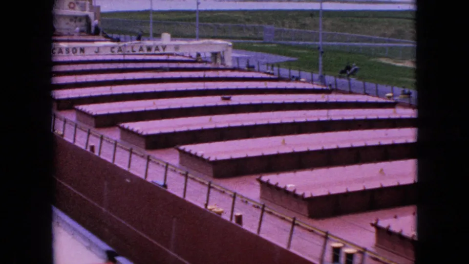 A closer 1964 home-movie view looks across the freighter's long deck, showing repeated cargo hatch covers, railings, partial ship name, and dockside grass beyond.