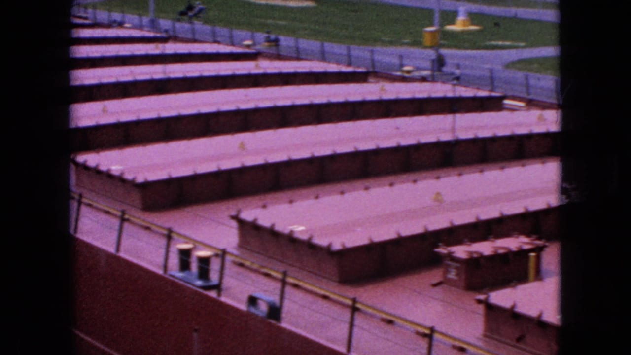 1964 Niagara Falls 8mm frame showing low reddish industrial rooftops in rows, with metal railings, paved surfaces, and grass visible beyond the facility.
