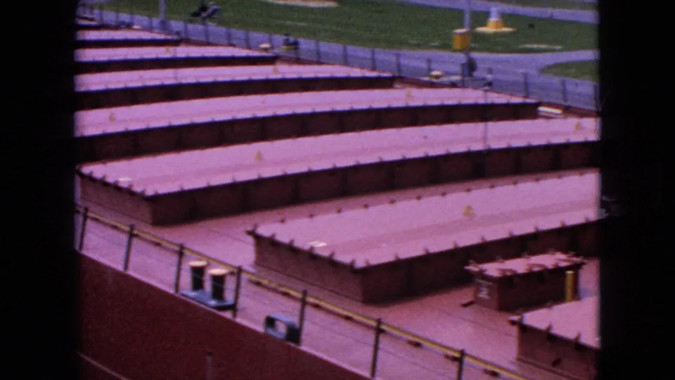 1964 Niagara Falls 8mm frame showing low reddish industrial rooftops in rows, with metal railings, paved surfaces, and grass visible beyond the facility.