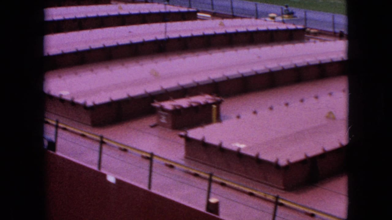High-angle 1964 Niagara Falls frame looking across a cluster of low industrial roofs, with railings, roof seams, small fixtures, and paved areas visible.