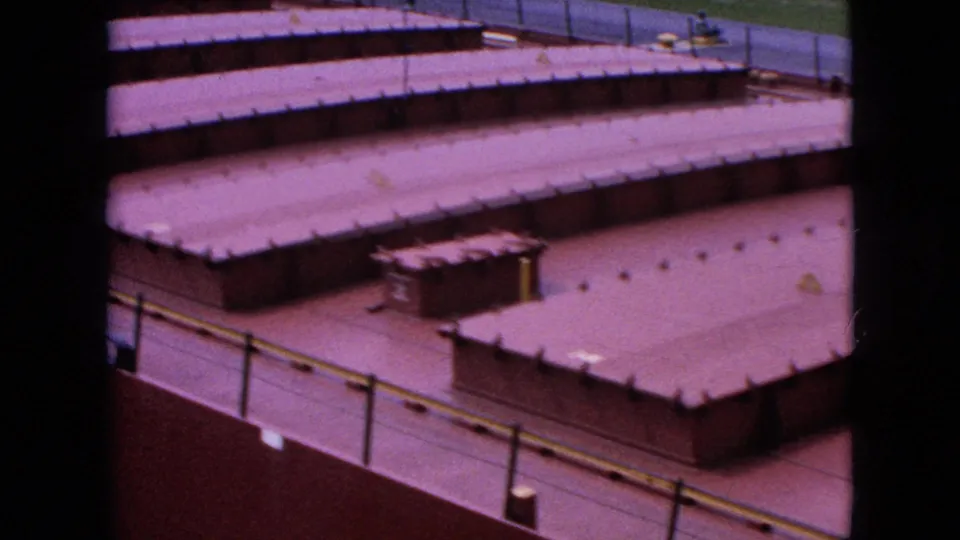High-angle 1964 Niagara Falls frame looking across a cluster of low industrial roofs, with railings, roof seams, small fixtures, and paved areas visible.