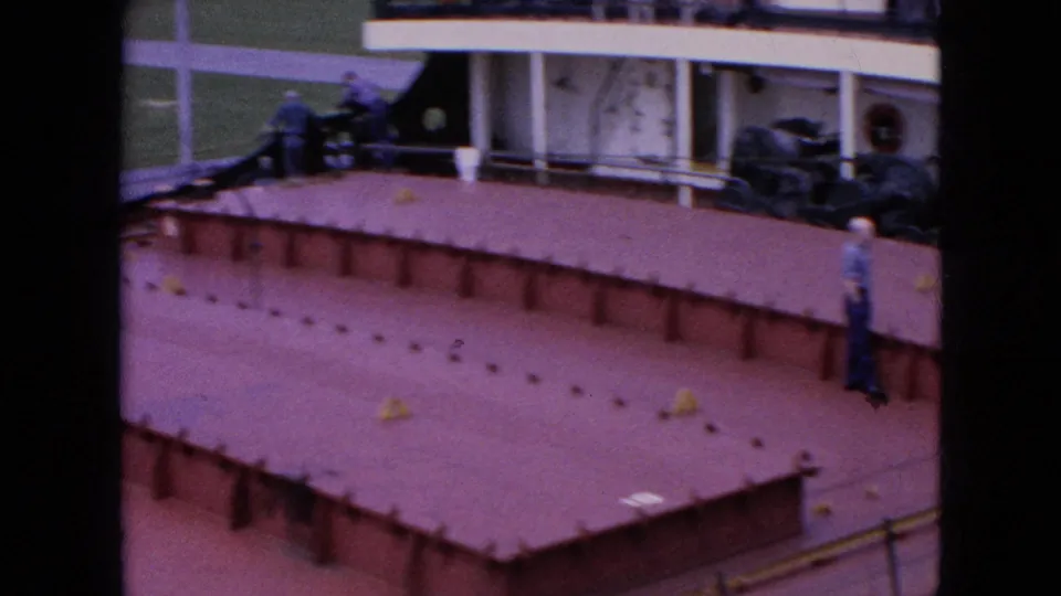 1964 home-movie frame showing crew members on or near a red tanker deck beside a white superstructure with railings, portholes, and deck equipment.