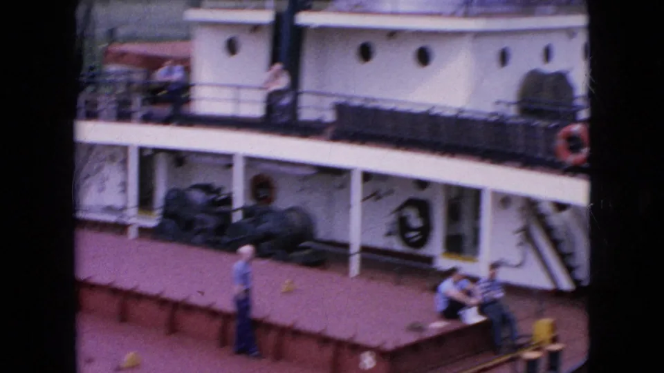 1964 8mm view of three ship workers around a red-painted tanker deck, with a white accommodation structure, railings, round portholes, and deck gear behind them.