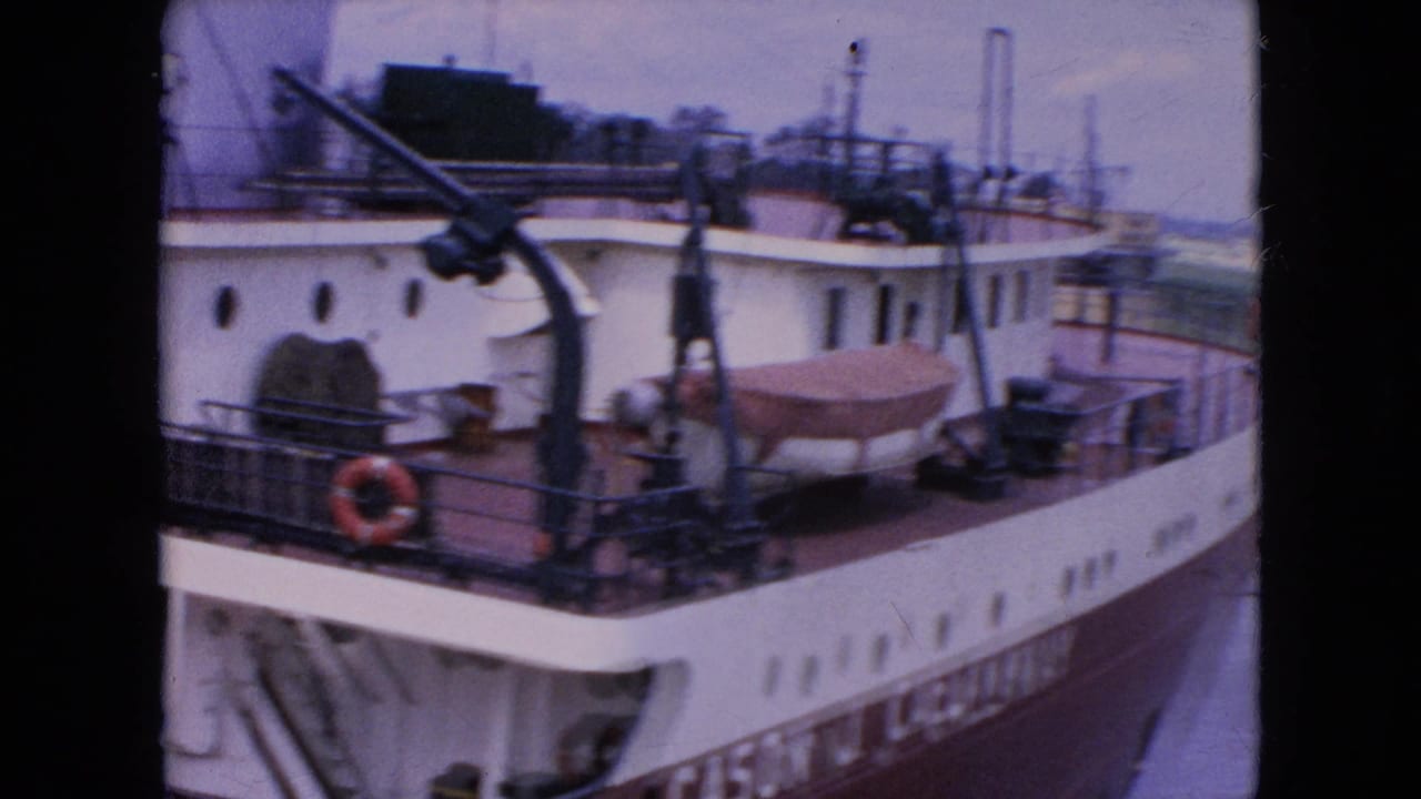 The Cason J. Callaway appears alongside a waterway or lock, with upper-deck cranes, railings, a red life ring, and a covered small boat visible in the 1964 home-movie frame.