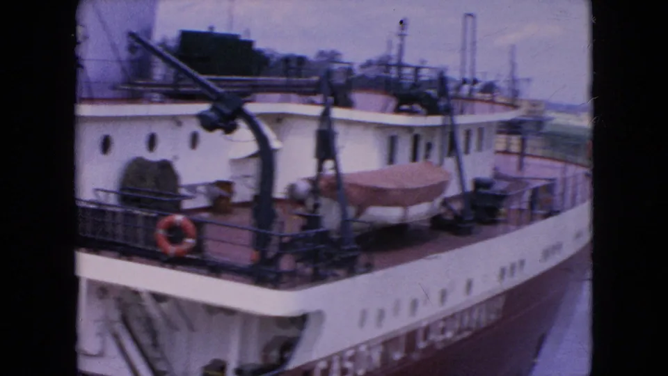 The Cason J. Callaway appears alongside a waterway or lock, with upper-deck cranes, railings, a red life ring, and a covered small boat visible in the 1964 home-movie frame.