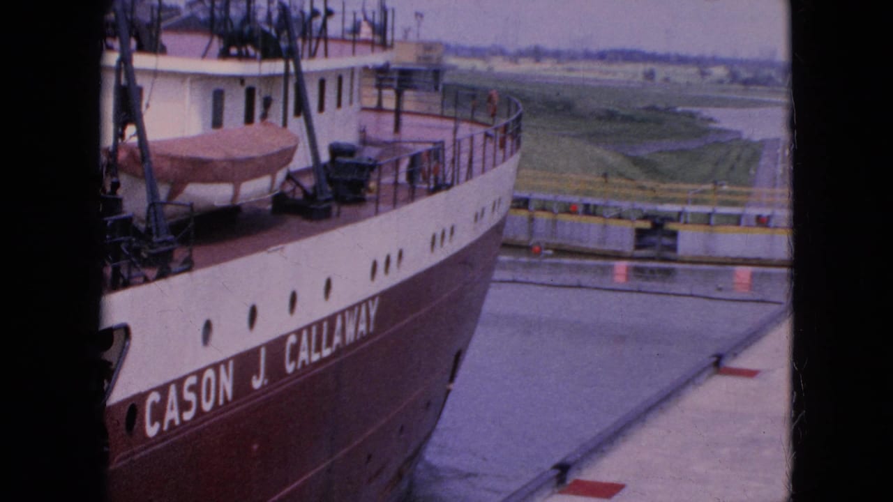 1964 8mm frame from Niagara Falls, New York showing the Cason J. Callaway freighter close to a lock wall, with deck equipment, a covered lifeboat, water, and flat land beyond.
