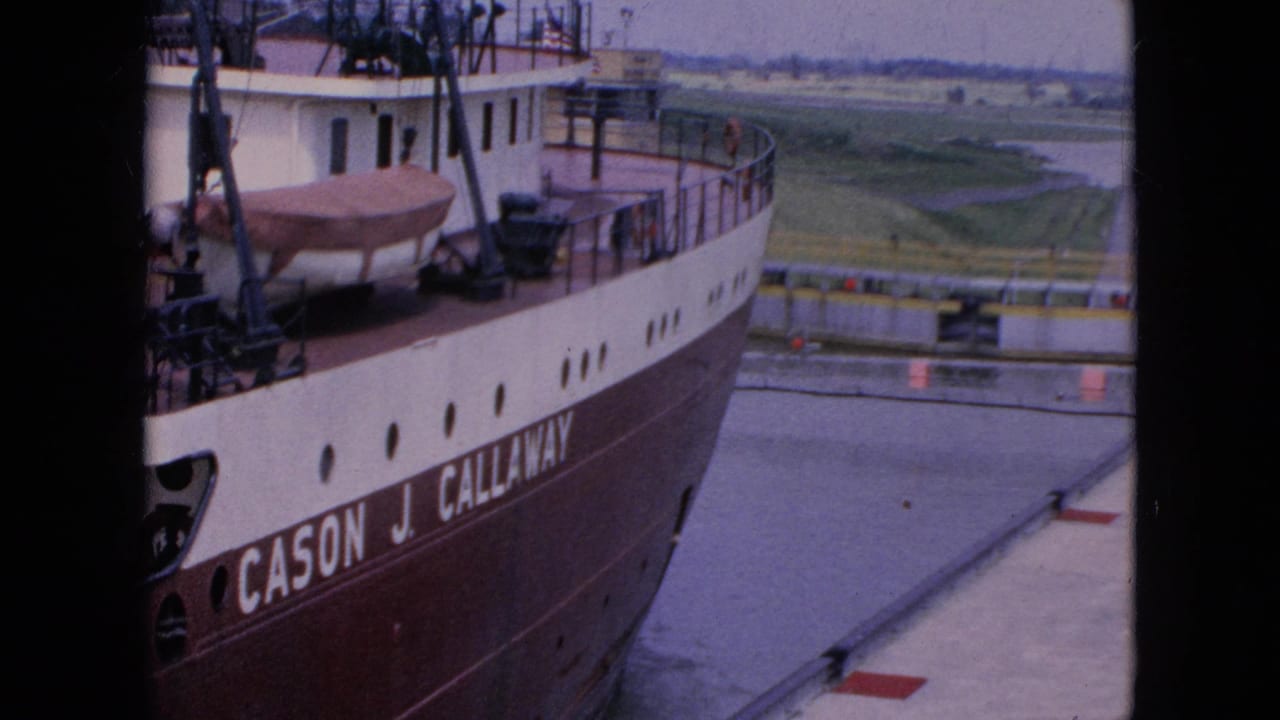Freighter bow and hull lettering beside lock water, with railings and low landscape in the distance.