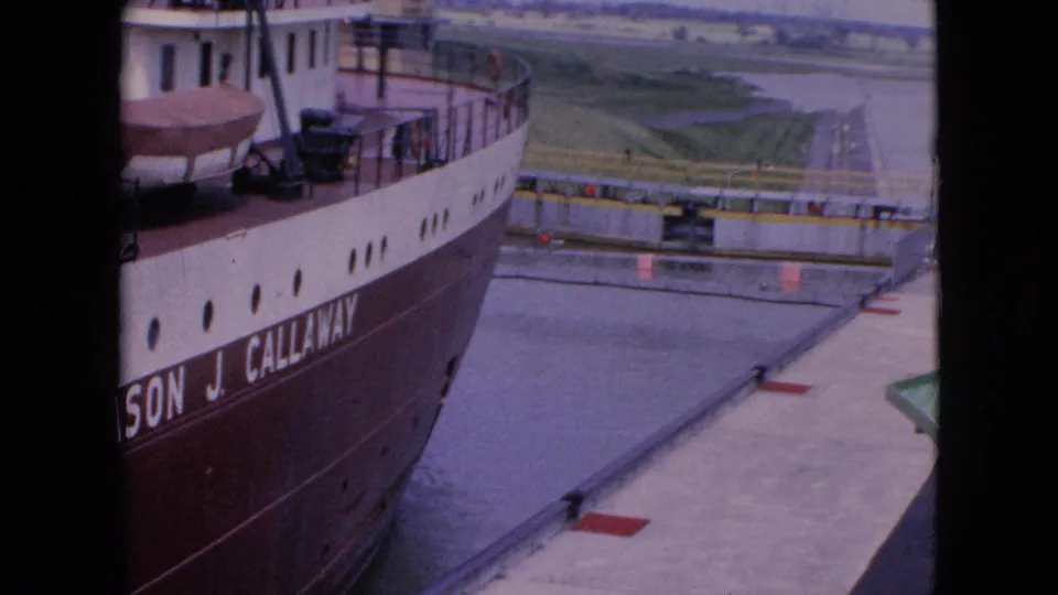 The freighter continues through the Niagara Falls, New York lock in a soft 1964 8mm frame, with partial Cason J. Callaway lettering, lock water, concrete margins, and distant flat terrain visible.