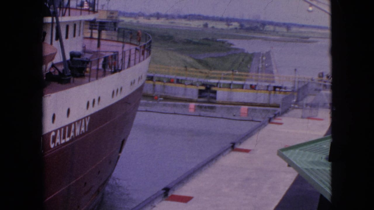 1964 8mm frame near Niagara Falls, New York, showing the side of a large freighter beside a lock wall with water and low green shoreline in the distance.