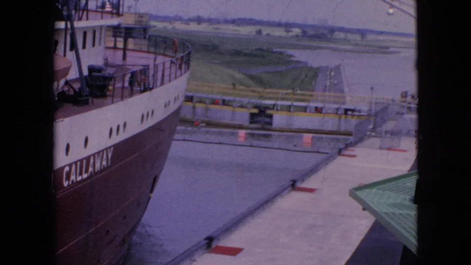 1964 8mm frame near Niagara Falls, New York, showing the side of a large freighter beside a lock wall with water and low green shoreline in the distance.