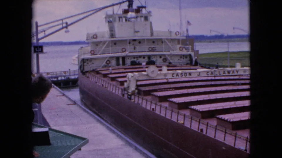 1964 8mm frame near Niagara Falls, New York, showing the Cason J. Callaway freighter aligned in a lock with repeated cargo-hold covers and a partly visible spectator in the foreground.