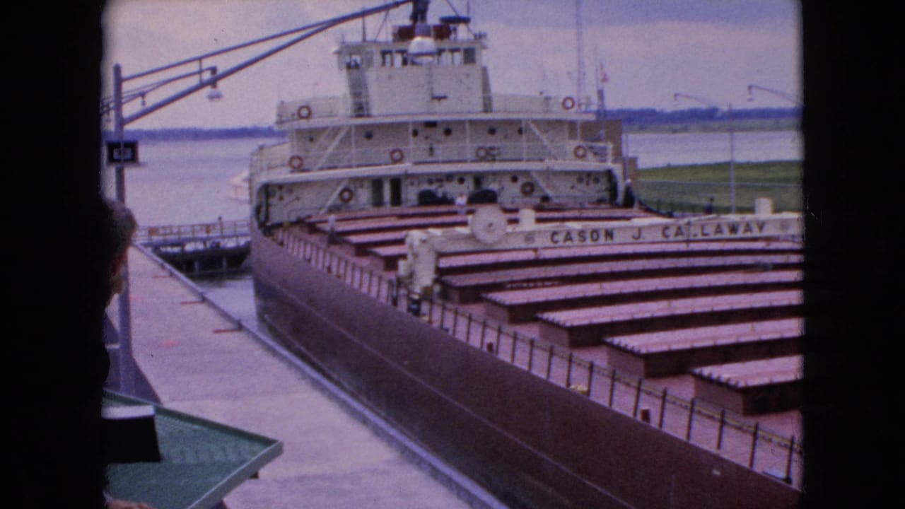 1964 8mm frame of the Cason J. Callaway docked at a U.S. waterfront, seen through a dark edge with cargo covers, superstructure, pier, and water visible.