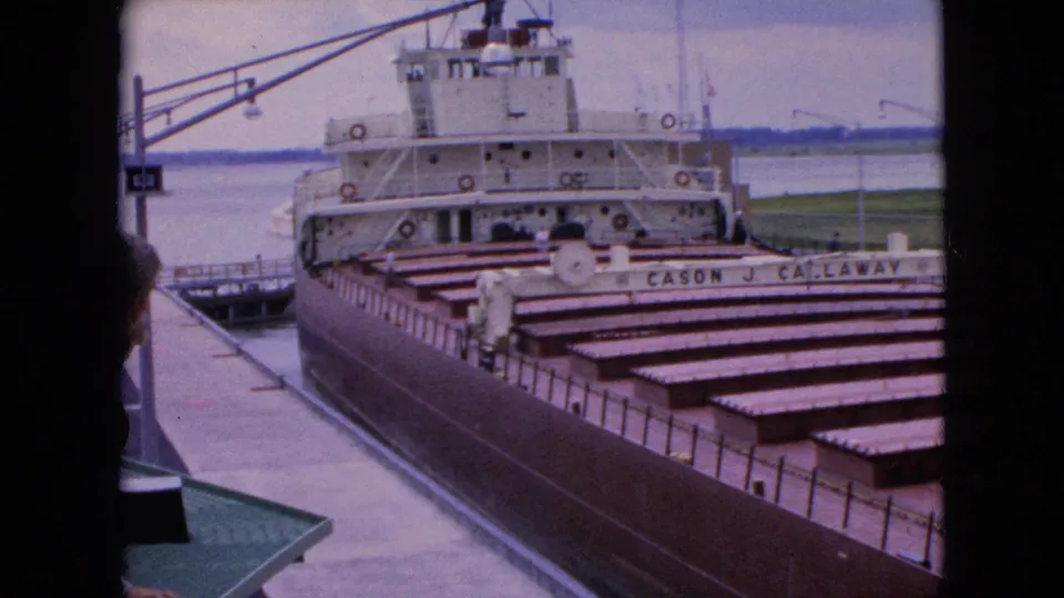 1964 8mm frame of the Cason J. Callaway docked at a U.S. waterfront, seen through a dark edge with cargo covers, superstructure, pier, and water visible.