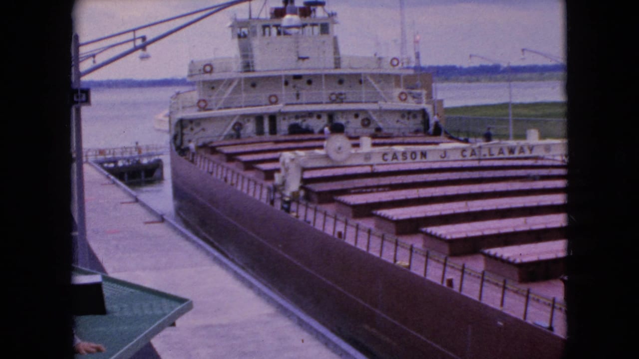 1964 8mm frame of the Cason J. Callaway docked near a pier, showing empty cargo holds, the white superstructure, water, and a distant shoreline.