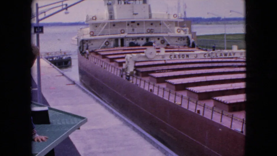 A slightly shifted 1964 8mm view shows the docked Cason J. Callaway, its open holds, white upper decks, nearby pier, water, and distant land.