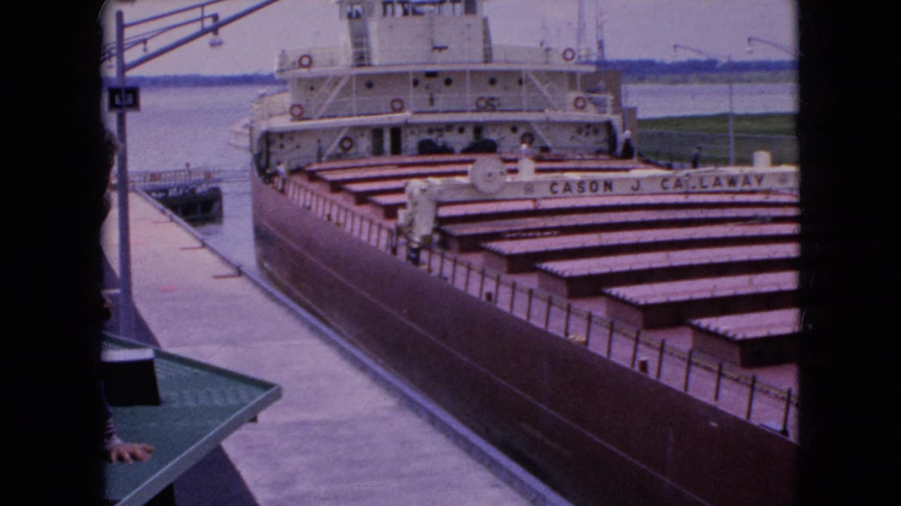 1964 United States 8mm frame showing the brown-hulled CASON J. CALLAWAY freighter moving beside a concrete dock, with a partial spectator visible near the camera.