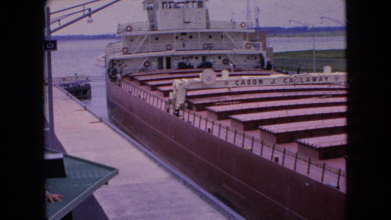 1964 United States home-movie frame of the CASON J. CALLAWAY freighter beside a dock, showing hatch covers, white deck structures, and nearby waterway.