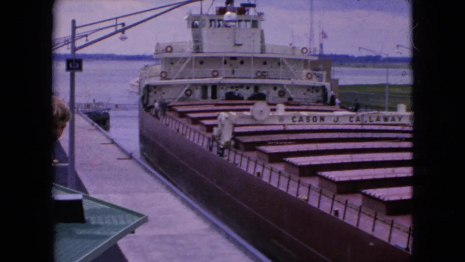 1964 United States home-movie frame of the CASON J. CALLAWAY passing a concrete dock, with cargo hatch covers filling the foreground and a partial onlooker at the frame edge.
