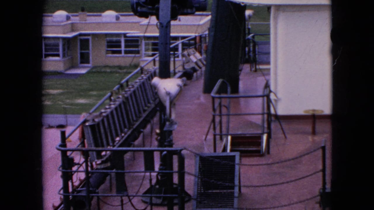 1964 8mm frame looking across shipboard railings, ropes, and deck equipment toward low dockside buildings near the waterfront.