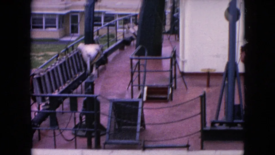 1964 home-movie frame showing deck railings, cable lines, a small ladder or gangway element, and a low dockside building beyond the vessel.