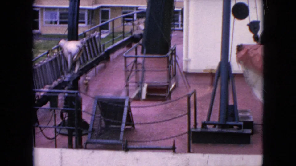1964 8mm frame of shipboard railings, cables, a deck hatch, and waterfront structures, with a partial person visible along the right edge of the frame.