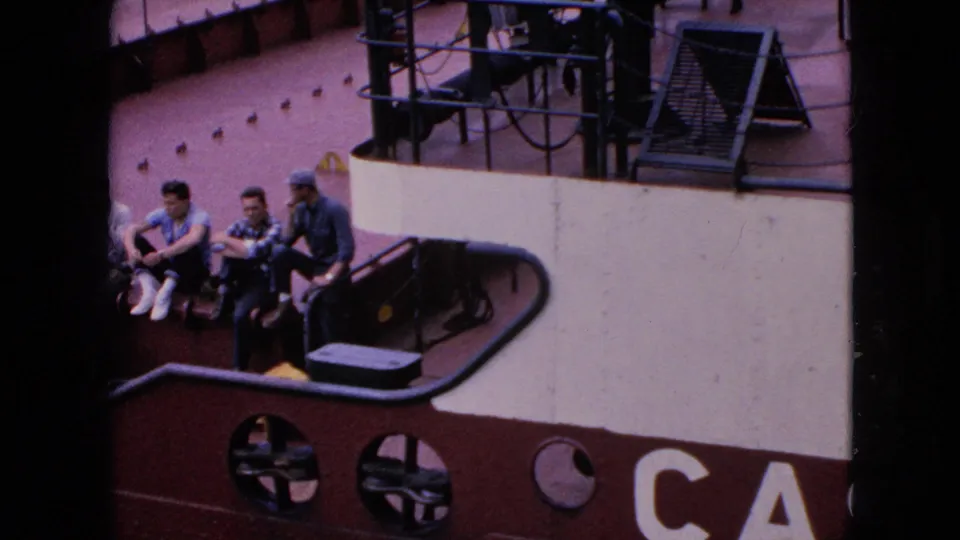 1964 Niagara Falls, New York home-movie frame showing three adult men in casual clothing sitting together on a red-and-white boat, with round portholes and railings visible nearby.