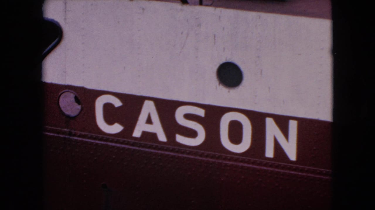 Close 1964 8mm frame of white block letters reading “CASON” on a burgundy band beneath a white painted metal panel, with dark round fittings visible.