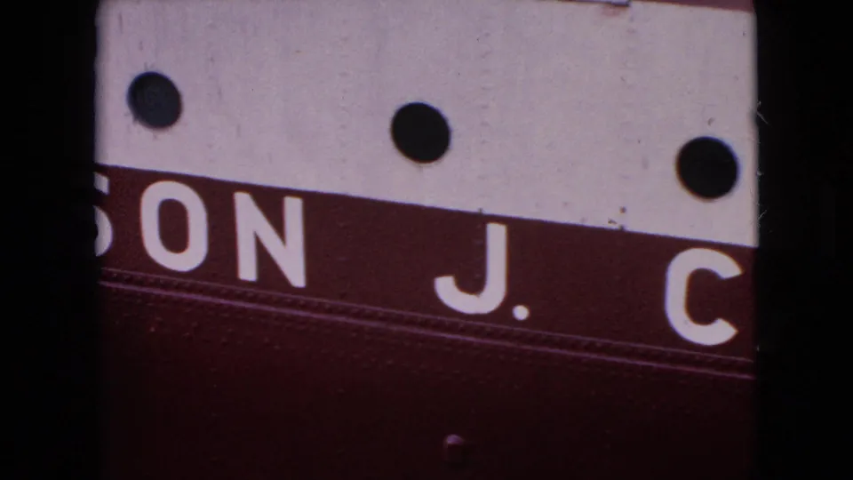 Close 1964 8mm frame of a cropped burgundy-and-white painted surface where the vessel name is partly visible as “ON J. C,” with round fittings above.