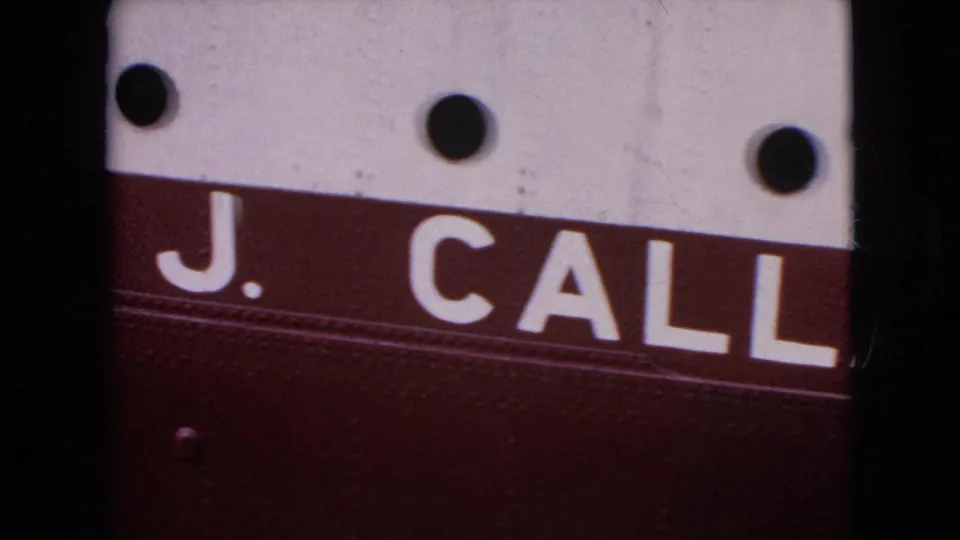 Close 1964 8mm frame showing the name segment “J. CALL” in white letters on a burgundy stripe, with white painted metal above and dark circular fittings.