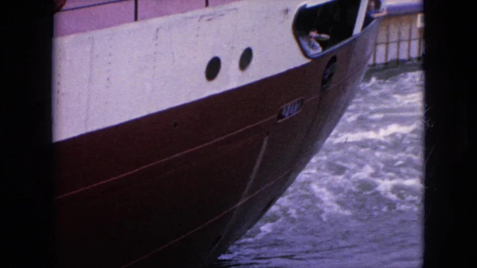 1964 8mm frame from the United States showing a low-angle view along the red and white boat hull with portholes and churning water beside it.