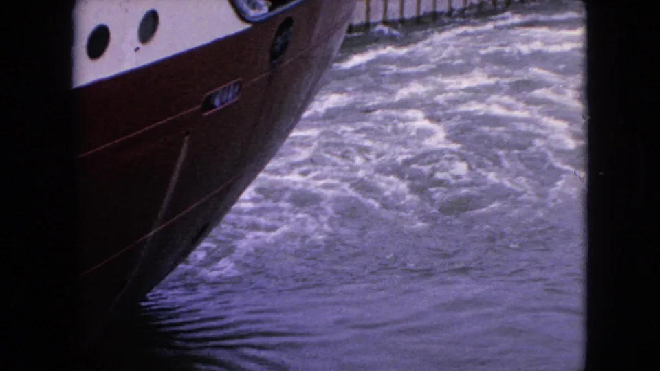 1964 8mm frame showing the boat's red lower hull and white upper hull moving through foamy wake and rippled water from a close side angle.