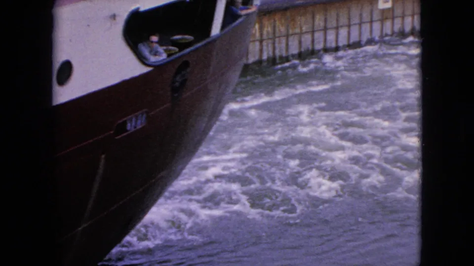 Red and white boat moving through foamy water near a wooden lock or channel wall, with a small person visible in the cabin area.