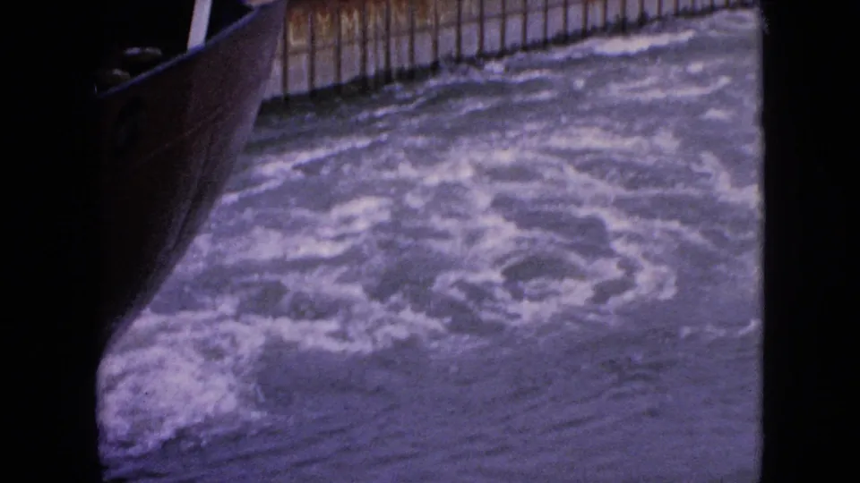 Close 8mm view of the boat edge and swirling water, with the wooden channel wall visible in the background.