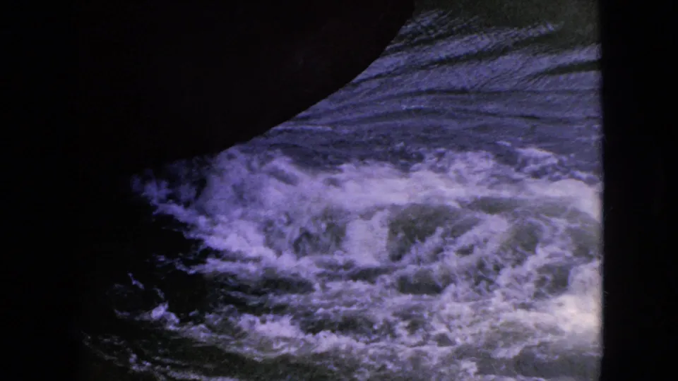 Shadowed boat bow crossing over turbulent, foaming water in color-shifted 8mm home-movie footage.