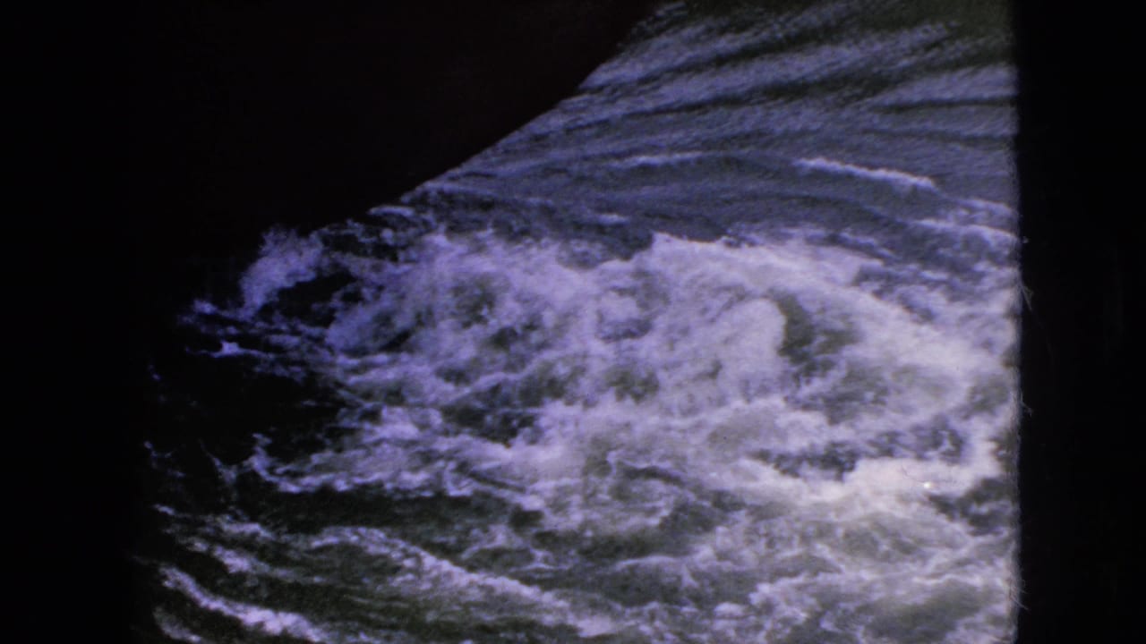 Turbulent water at a waterways setting, likely from the Niagara Falls travel footage, filmed through a dark structural or window-like frame on 8mm film.