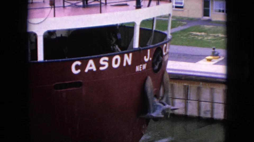 A red-hulled vessel with partial white lettering appears docked beside a canal lock wall, with deck railings and nearby building details visible in the 1964 footage.