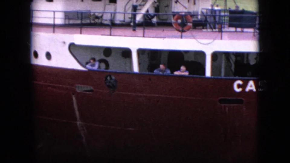 Passengers are visible through rectangular windows on a red-hulled boat near a dock or lockside, with partial vessel lettering and deck equipment in view.