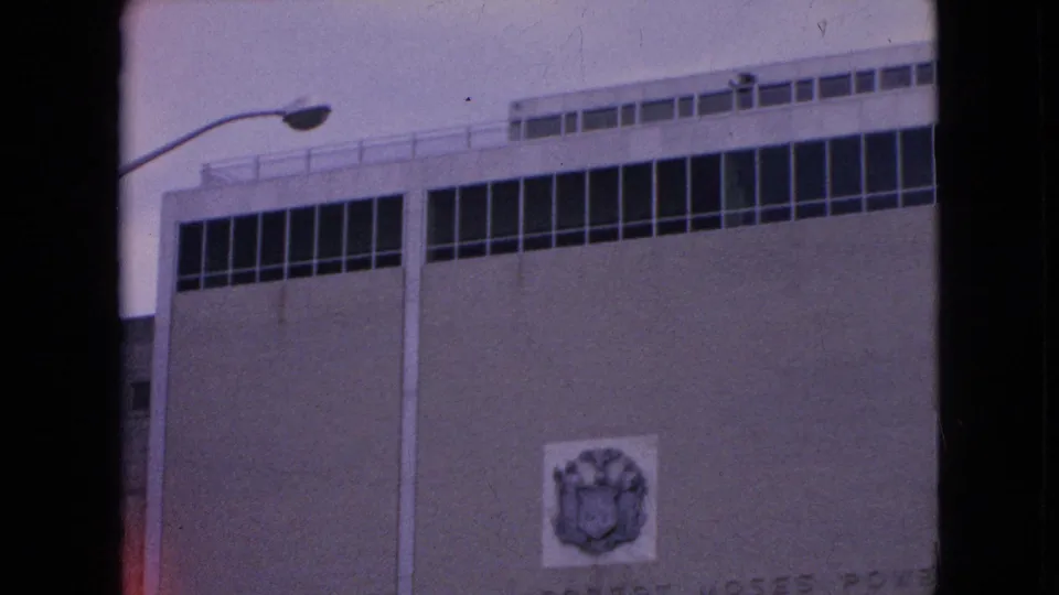 1964 8mm frame of a large building facade with rows of windows, a streetlight, and a Robert Moses Power Dam sign on the wall.