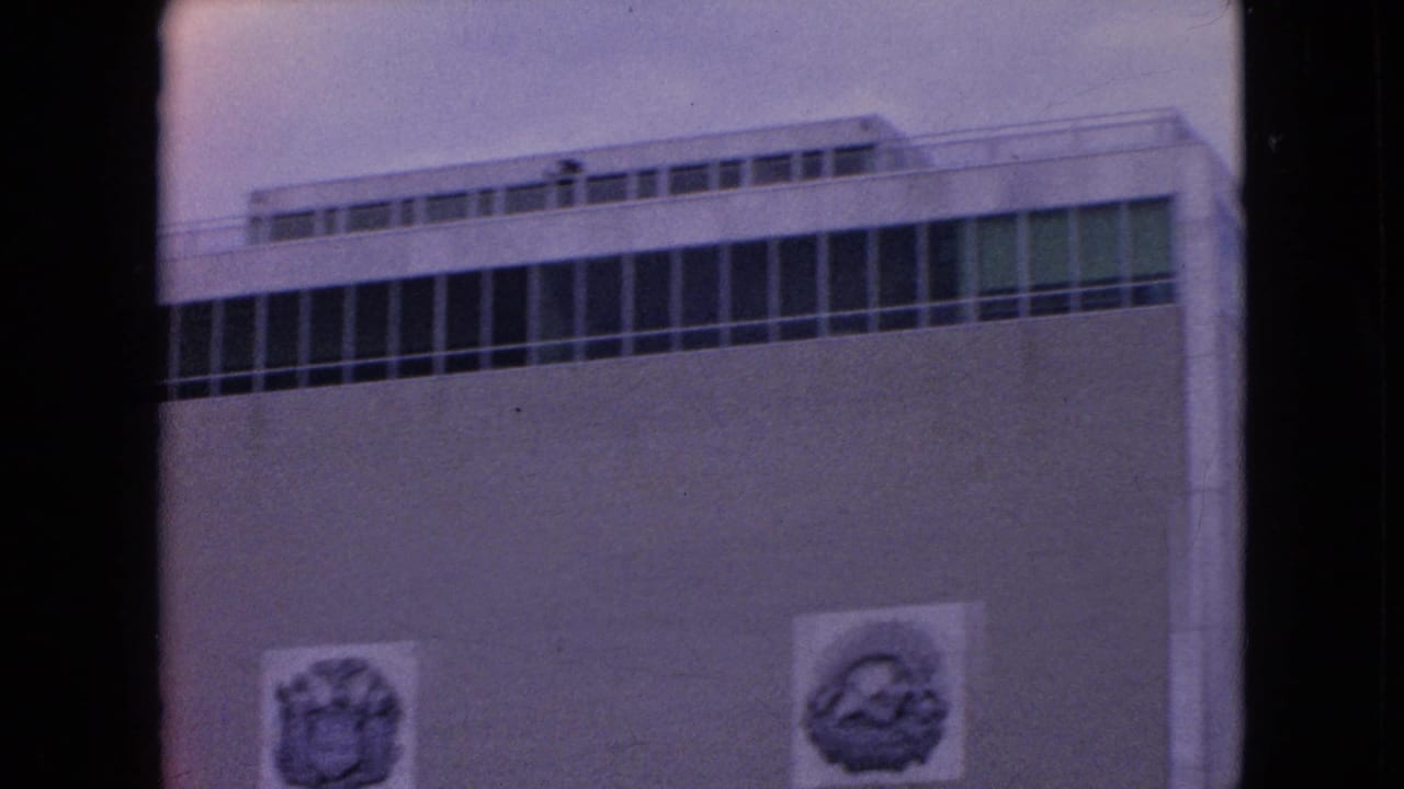 1964 8mm frame from Niagara Falls, New York showing a flat-roofed modernist building with upper window bands above a concrete dam wall and water.