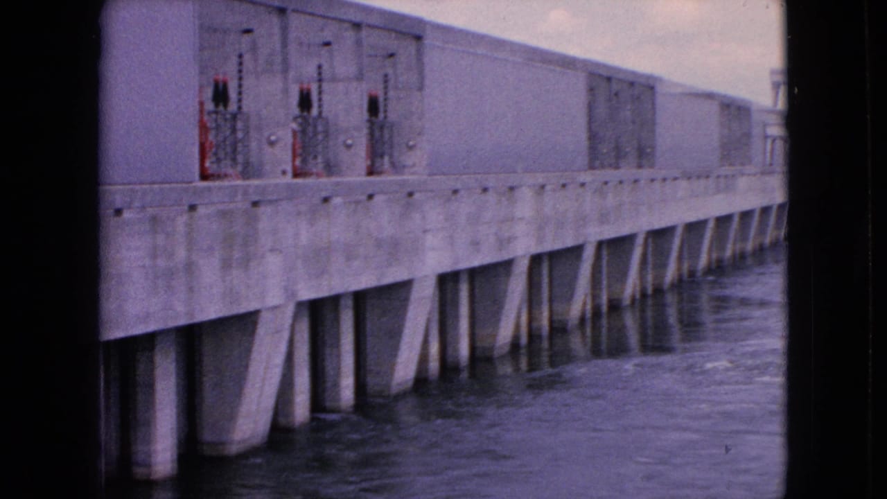 1964 8mm frame from Niagara Falls, New York, showing a long concrete hydro dam wall, metal control equipment, and water moving below the supports.