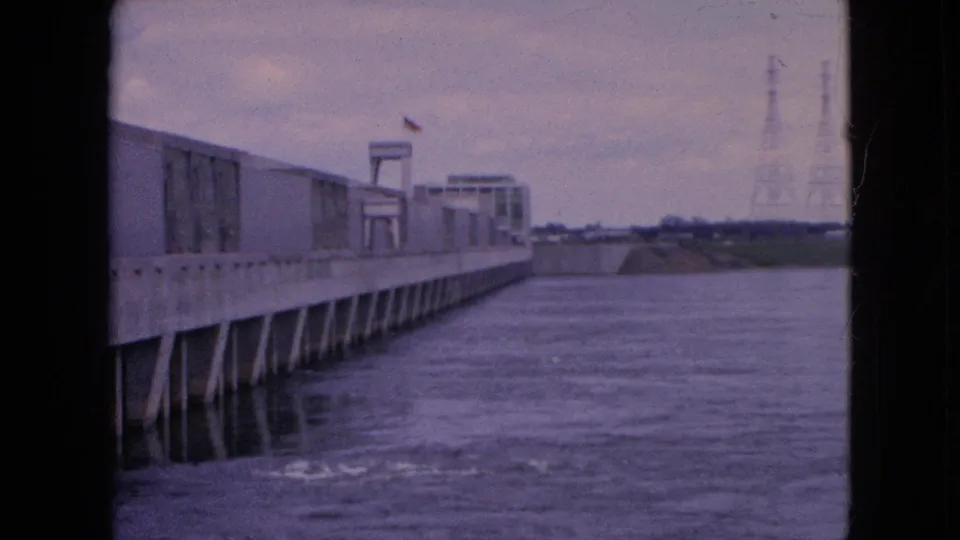 1964 8mm frame showing the Niagara Falls hydro dam from the side, with rippled water, concrete supports, distant tower structures, and a small flag visible.