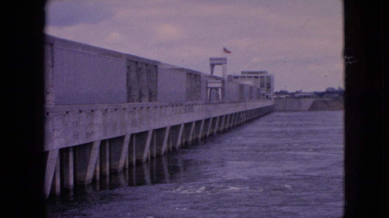 1964 home-movie frame from Niagara Falls, New York showing a long concrete dam or lock-like structure beside flowing river water, with a small flag near a tower form.