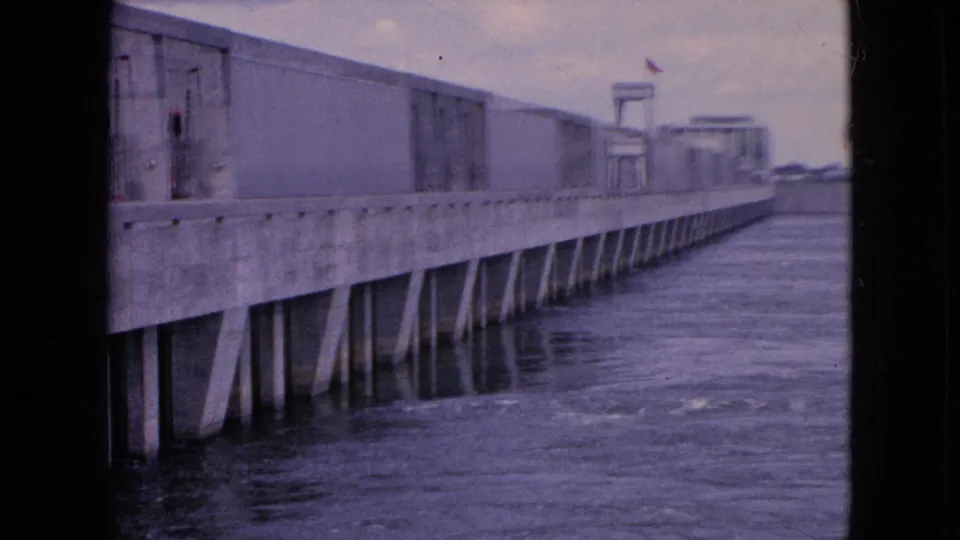 A slightly closer 1964 8mm view shows the concrete riverside structure passing by the camera, with vertical and angled supports reflected in the water.