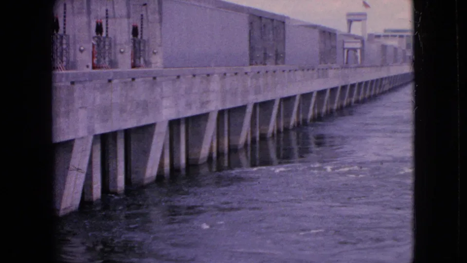 The final frame shows the concrete industrial wall from a closer angle, with dark fixtures or rail elements above the structure and river water moving alongside it.