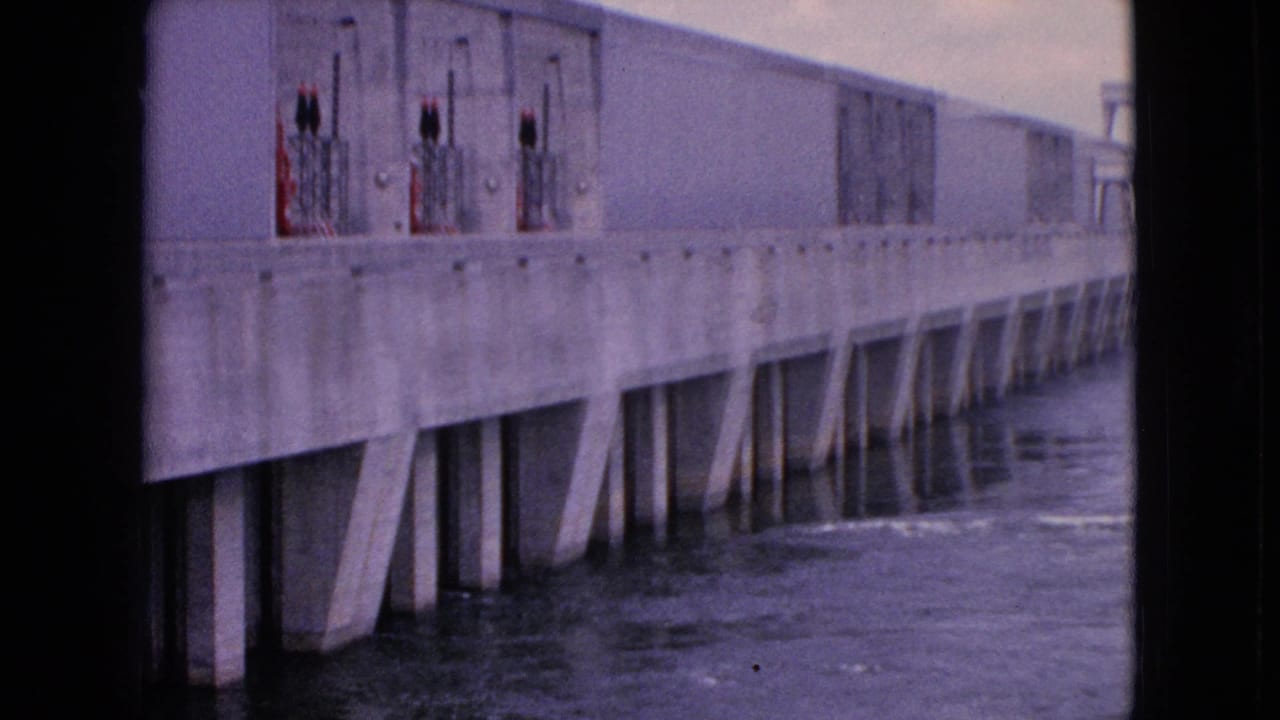 1964 8mm frame from Niagara Falls, New York showing a long concrete bridge or dam-like structure with angled supports extending into dark moving water.