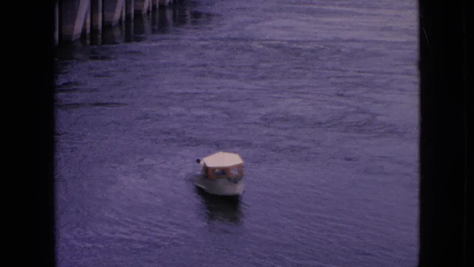 A small motorboat with a light canopy moves on the river beside the large concrete structure in this 1964 Niagara Falls, New York home-movie frame.