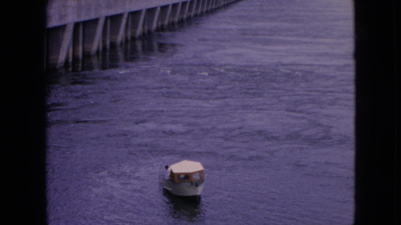 A small white covered motorboat appears on rippled water in 1964 Niagara Falls-area 8mm footage, with dark vertical frame edges and distant pilings visible.