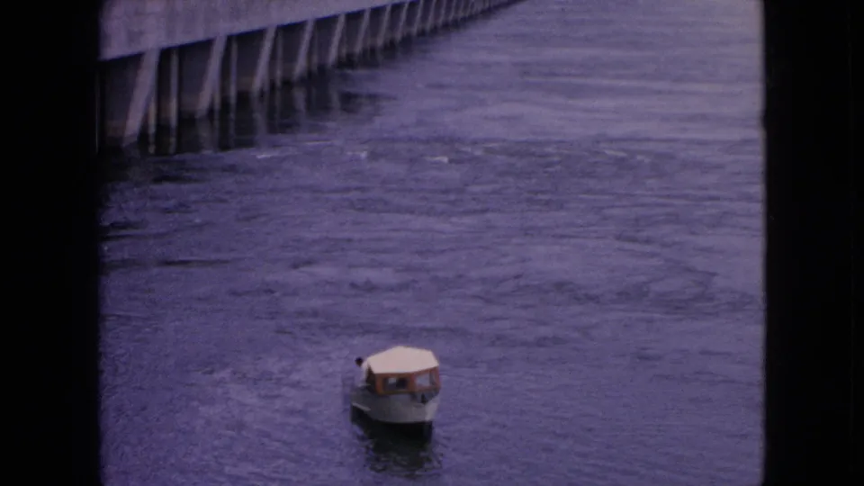 A small white covered motorboat appears on rippled water in 1964 Niagara Falls-area 8mm footage, with dark vertical frame edges and distant pilings visible.